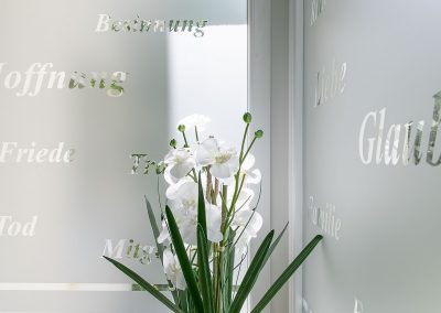Glaswand mit Trauersprüchen, davor eine Pflanze mit edlen, weißen Blüten